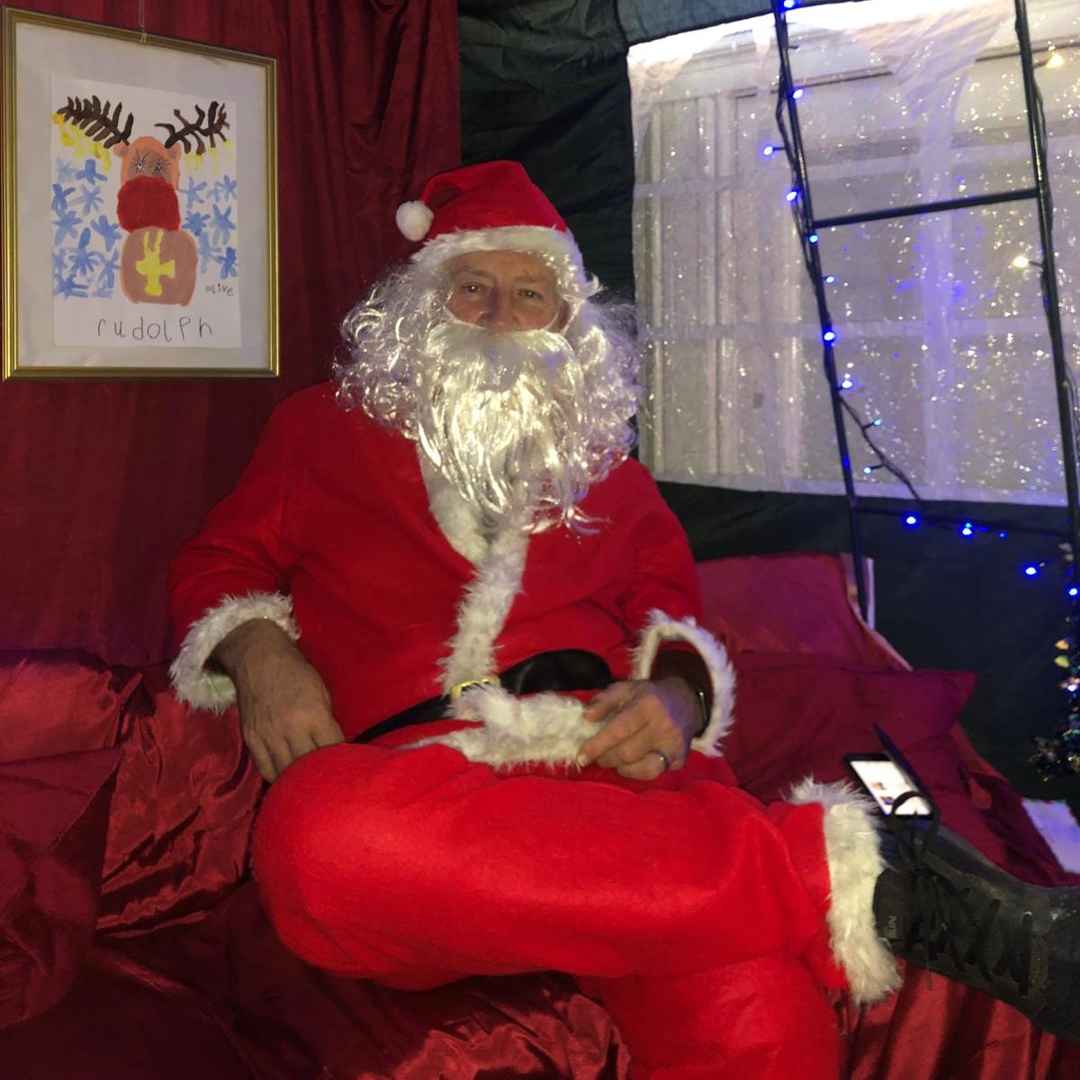 A man in a Santa Claus costume sits on a red couch, smiling cheerfully, embodying the festive spirit of Christmas.