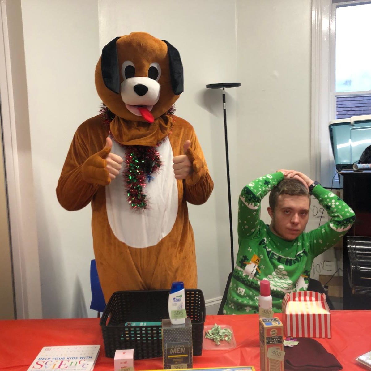 A man in a dog costume stands by a table with a book and various tombola items, creating a fun atmosphere.