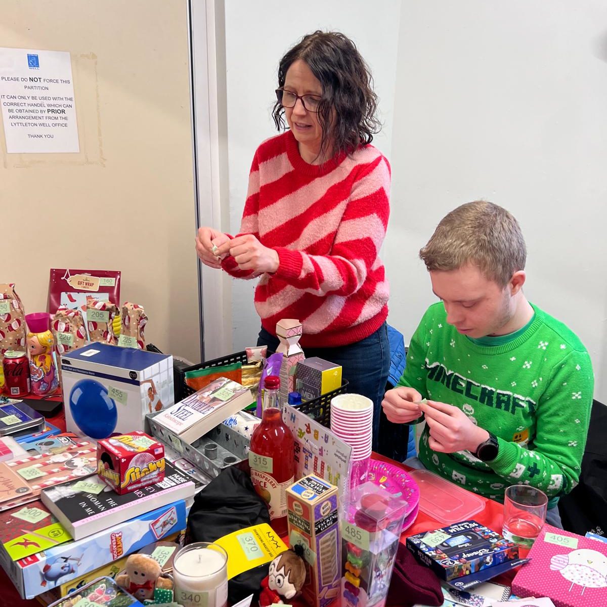  A woman and a boy are seated at a table, surrounded by various items, as they look at tombola tickets and run the stall.